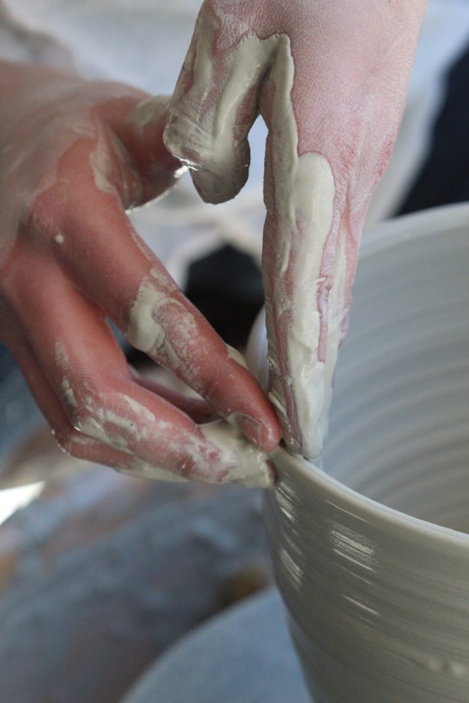 The fingers of a person with pale skin delicately manipulate wet clay on a pottery wheel. The hands are wet, covered with shiny clay.