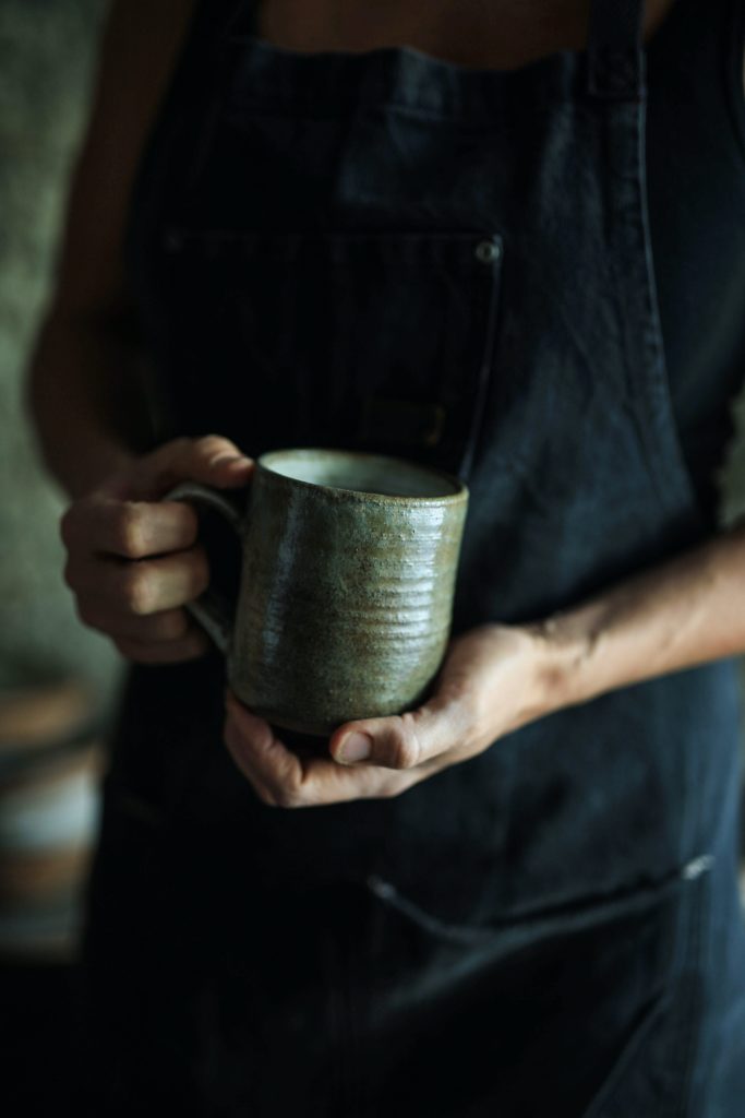 A person with pale skin in a black apron holds a handmade mug which is olive in color.