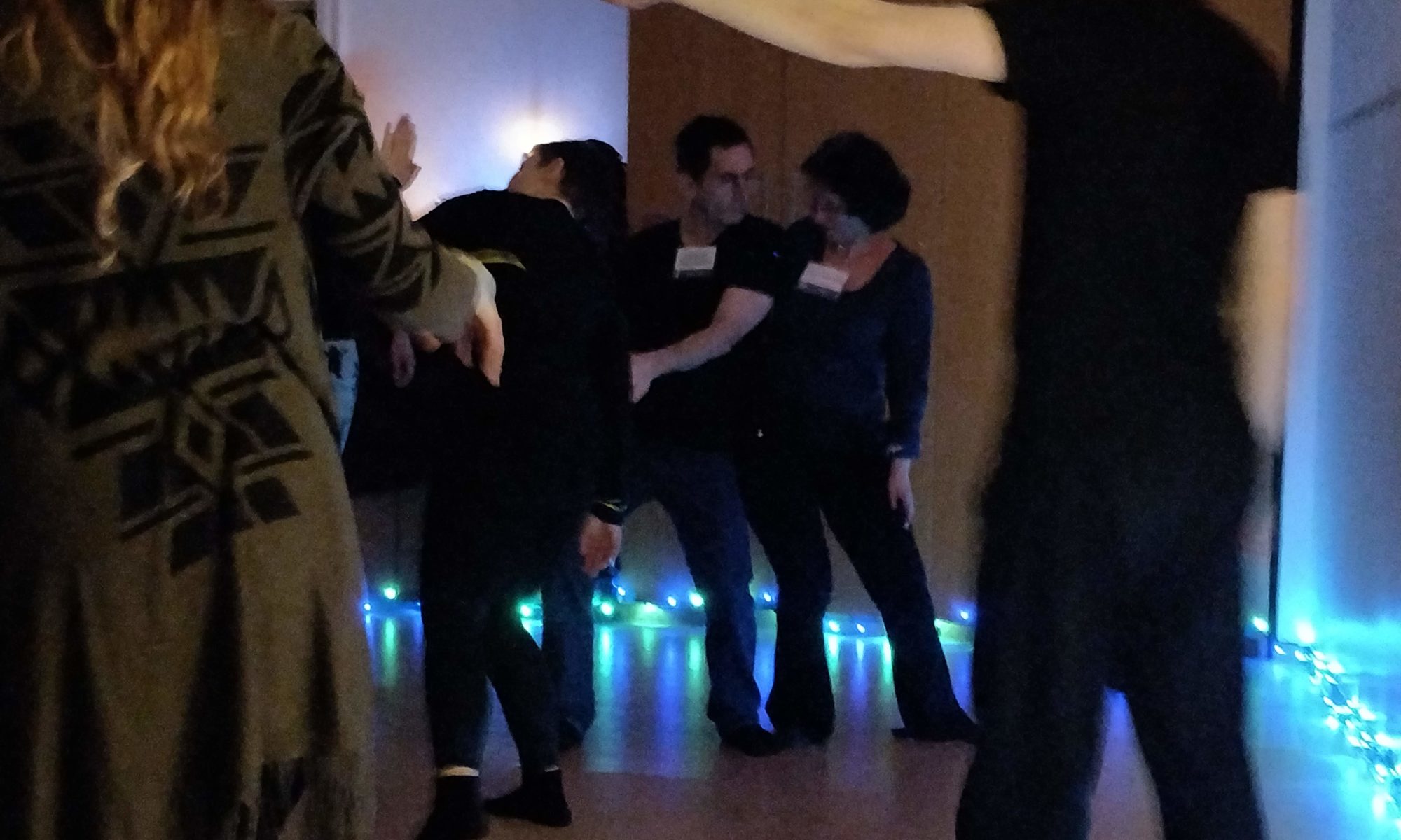 People dance in a dimly lit room. Two move lean into one another, sharing weight. One person the the foreground thrusts out an outstretched hand to another person.