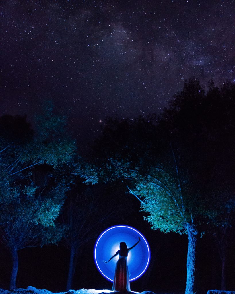 Woman in a forest at night, spinning her arms. A glowing blue circle of light appears behind her, illuminating the darkness.
