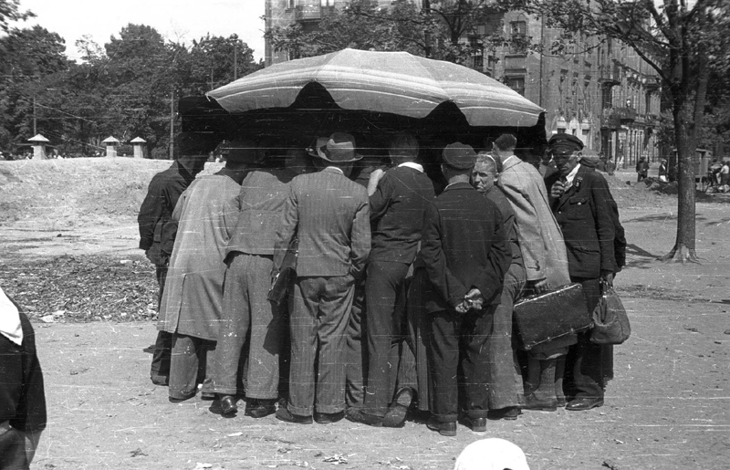 A black-and-white photo of people in 1940s clothing gathered around an umbrella covered stall