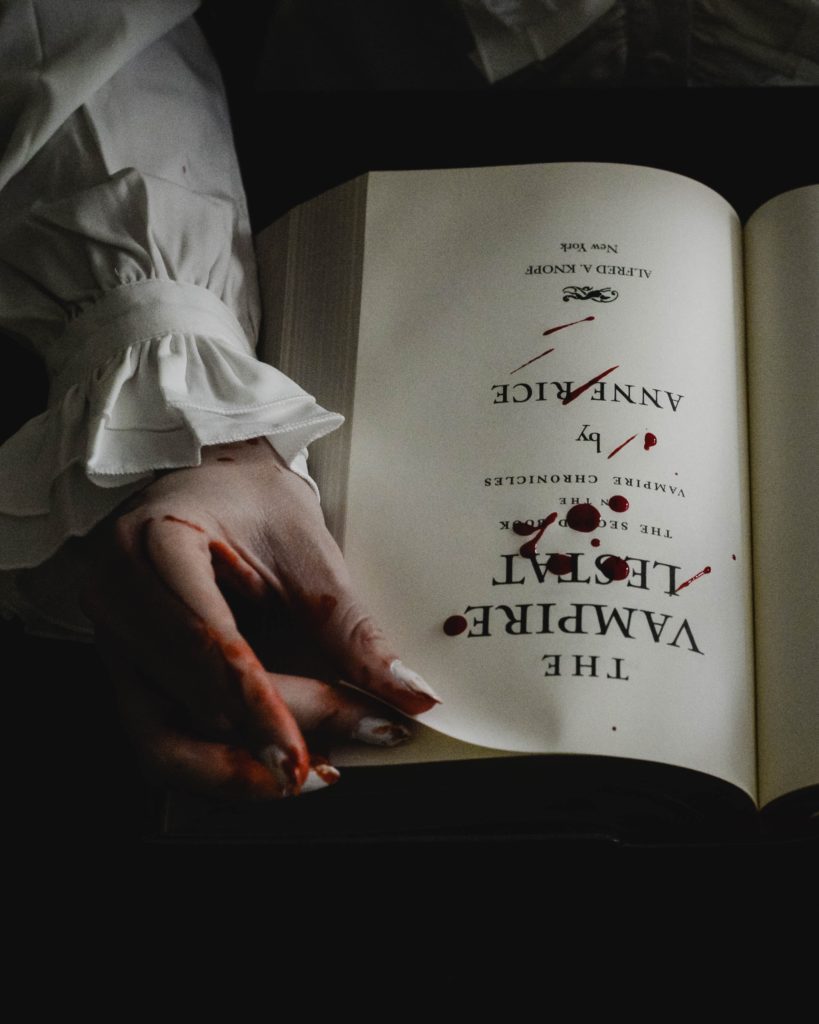 A bloody hand with a ruffled sleeve holds open the blood-spattered title page of The Vampire Lestat by Anne Rice