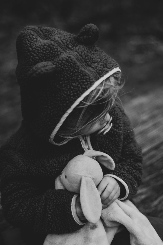 A little girl with light skin whose face is obscured by a hood clutches a stuffed rabbit to her chest, as if for comfort.