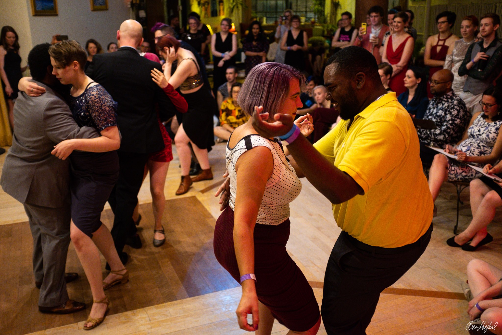 Multiple dancing duos compete at North Star Blues, in front of an audience and judges. One partnership is centered in the frame, the two dancers make eye contact while in closed position. The leading person is Black and masc, wearing a yellow polo and black pants. The person following is a white femme in a polkadot top and red skirt.