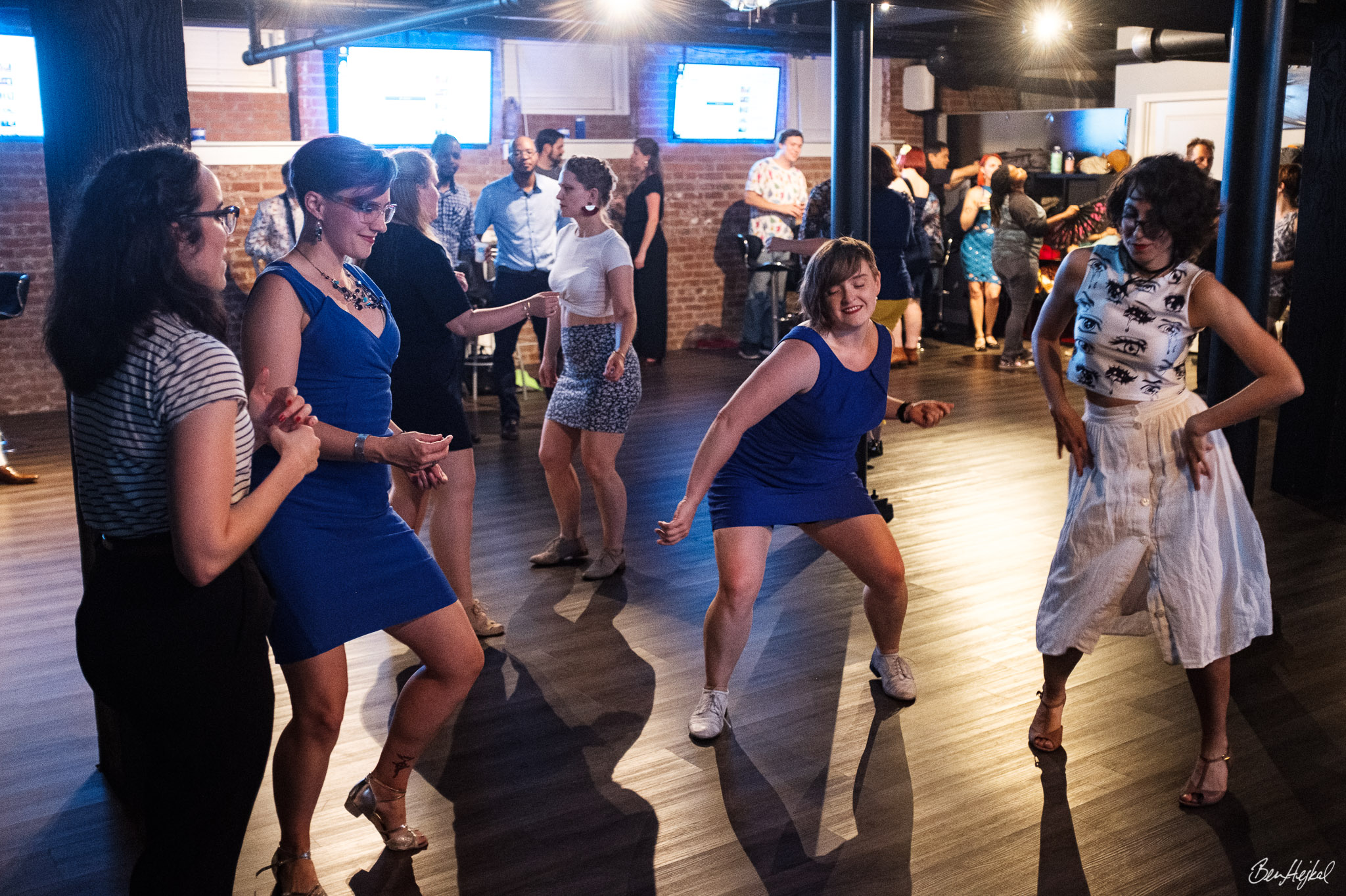 A full dance floor at a dance event. In the center of the frame four femmes dance solo, riffing on one another's moves.