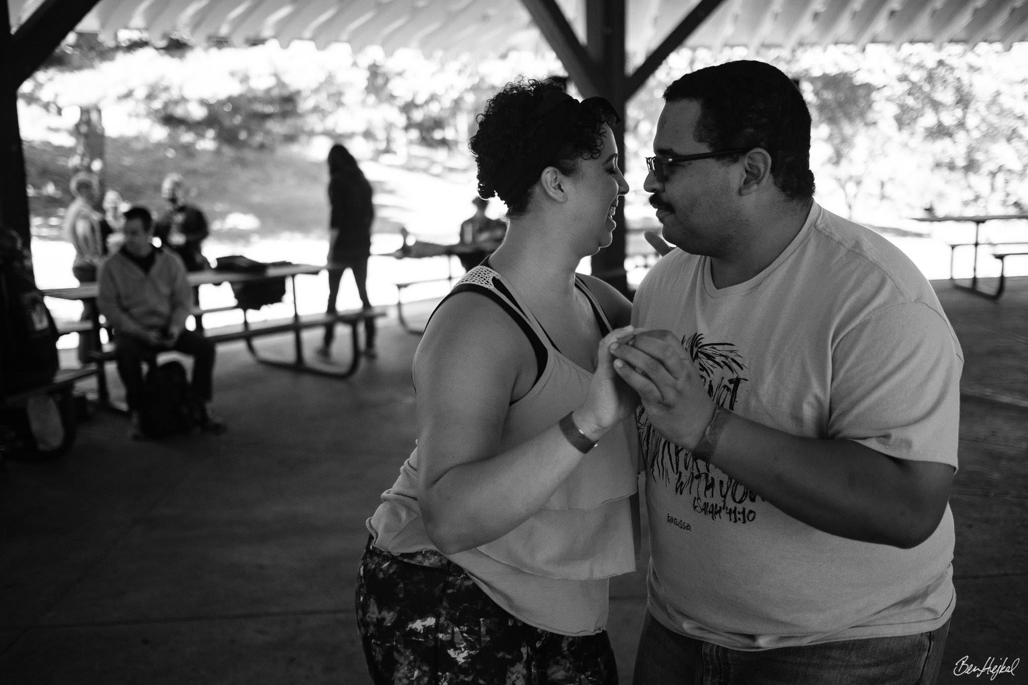 Two fat Black people dance in close embrace under a shady picnic pavilion at a park. The masc person leading has a mustache and glasses and is wearing a tshirt and jeans. The femme person following is smiling, wears a flowing top and leggings.
