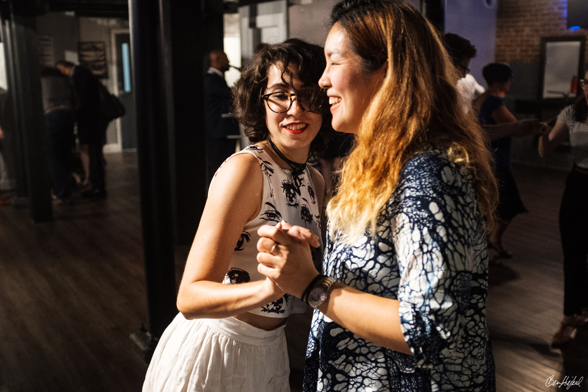 Two femmes dance in closed position at a dance event, surrounded by other dancers. The person leading is a AAPI femme with long dyed blonde hair in a patterned blouse. The person following is a white femme with dark bobbed hair in a patterned top and white skirt.