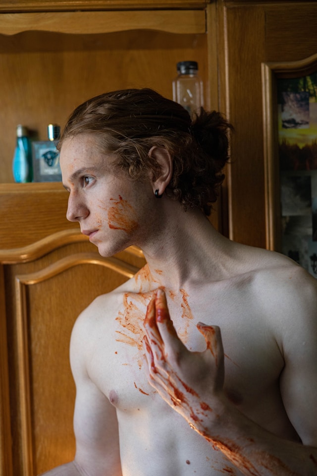 A shirtless young man with pale skin stares in profile. His raised hand is covered in blood and he seems to have carelessly wiped some onto his cheek and chest.