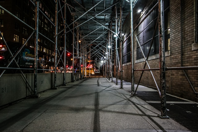A low angle photograph of a New York City sidewalk at night. The wide thoroughfare is narrowed by scaffolding, forming an empty tunnel.
