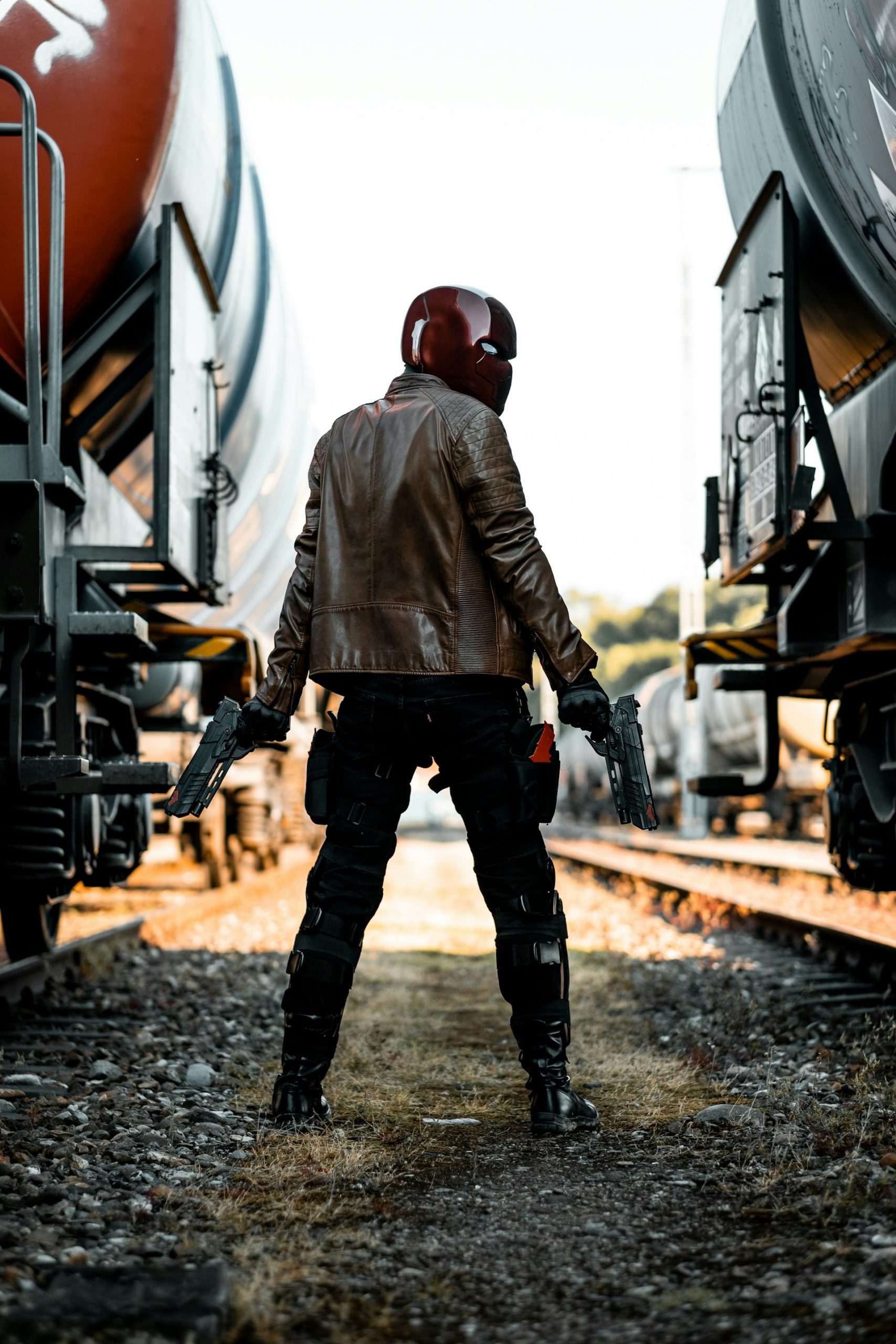 A masked villain stands facing away from the viewers, looking back over their shoulder. Flanked by 2 trains, they are holding 2 scifi guns and conceal their identity with a full coverage helmet.